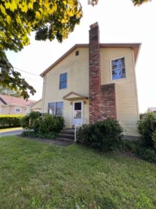 A yellow house with a brick chimney and bushes.